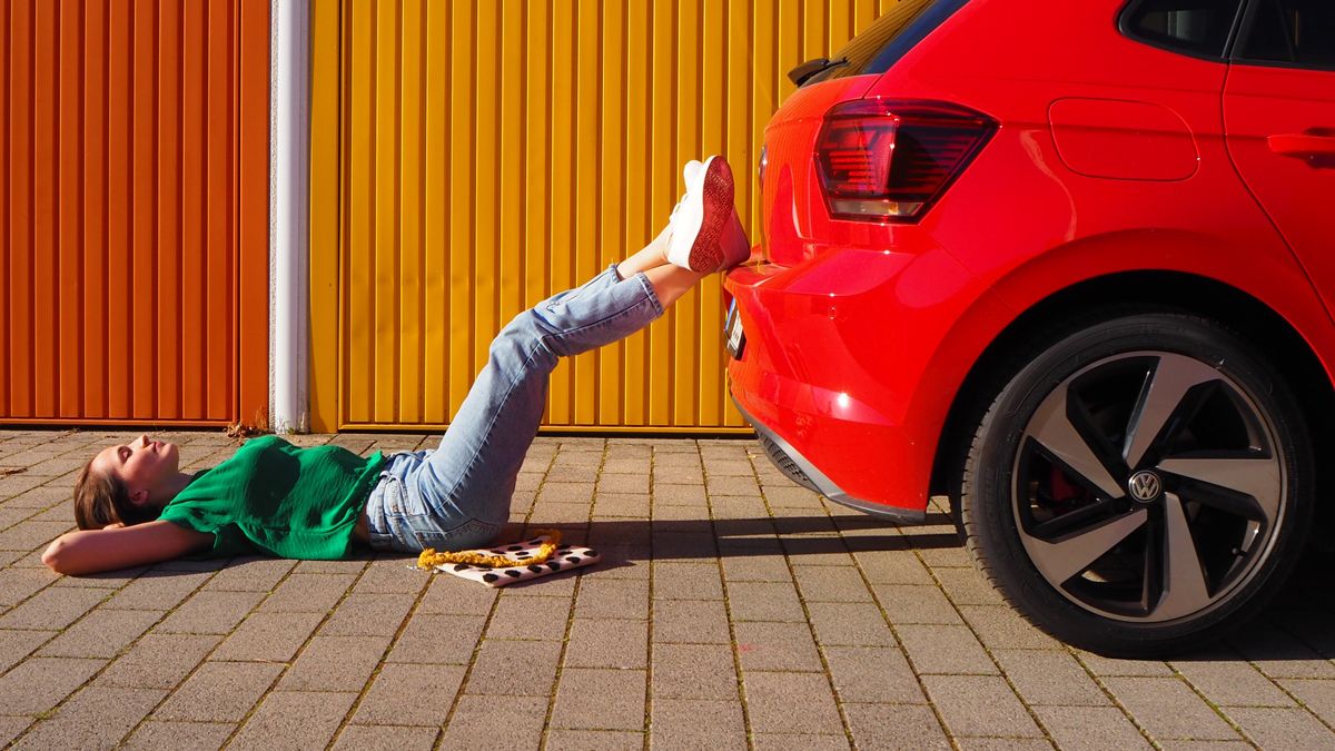 Euro NCAP | Safety assurance A casually dressed lady lying down and resting her legs on the bumper of a red Volkswagen vehicle parked beside an outdoor urban area under a sunny weather.