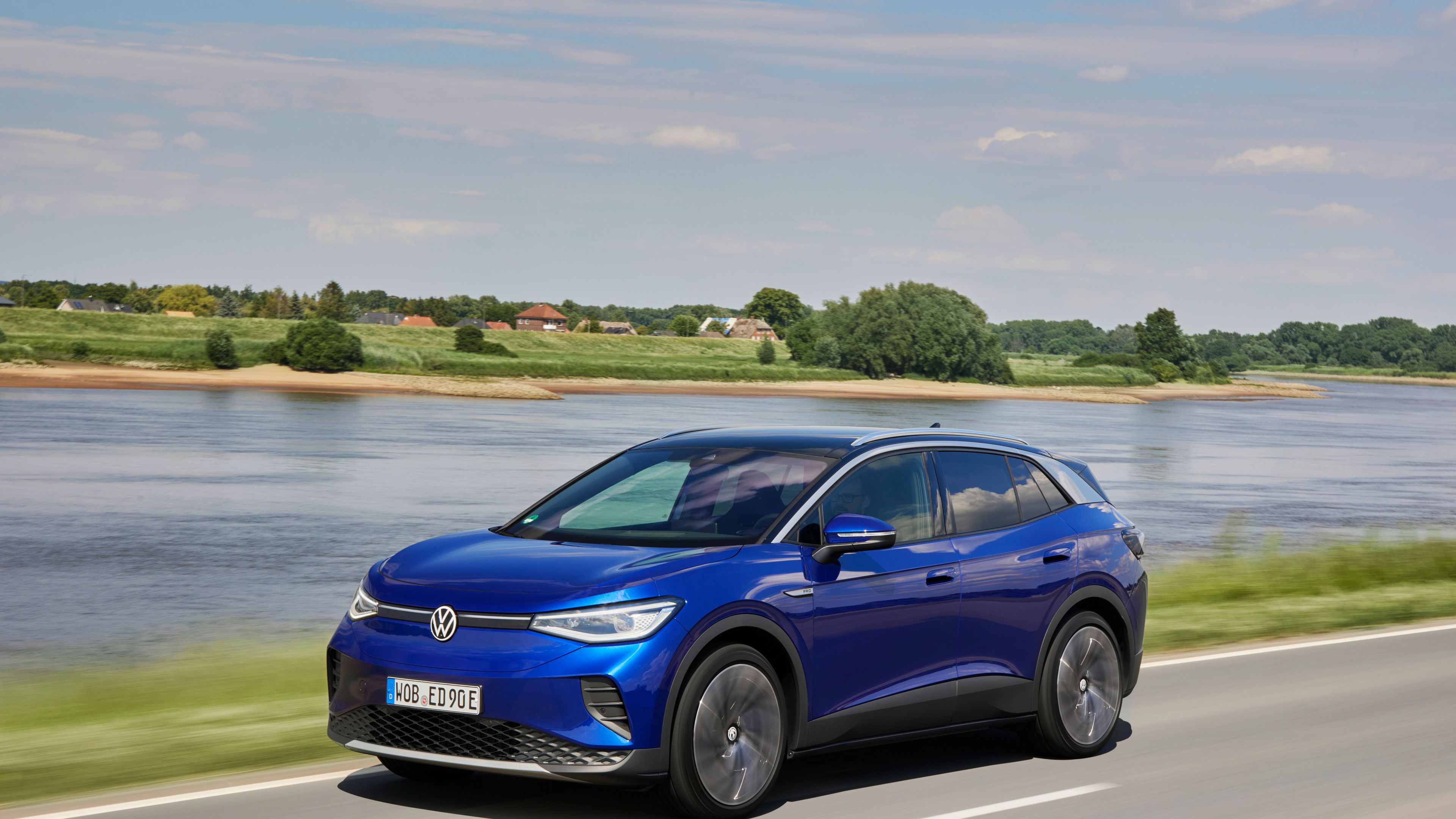 Volkswagen ID.4 A blue Volkswagen ID.4 driving down a road with a lake and lush greenery beside it